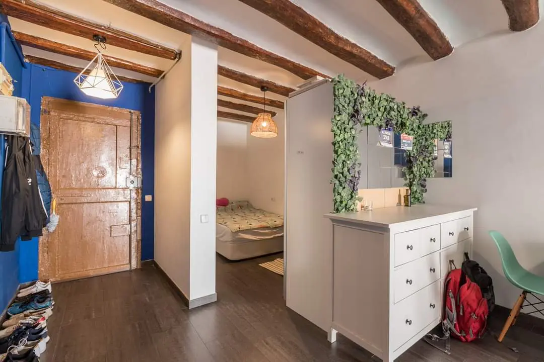 Bedroom interior with exposed beams, wood floors, and a blue accent wall. A bed is visible through a doorway. A white dresser is decorated with greenery.