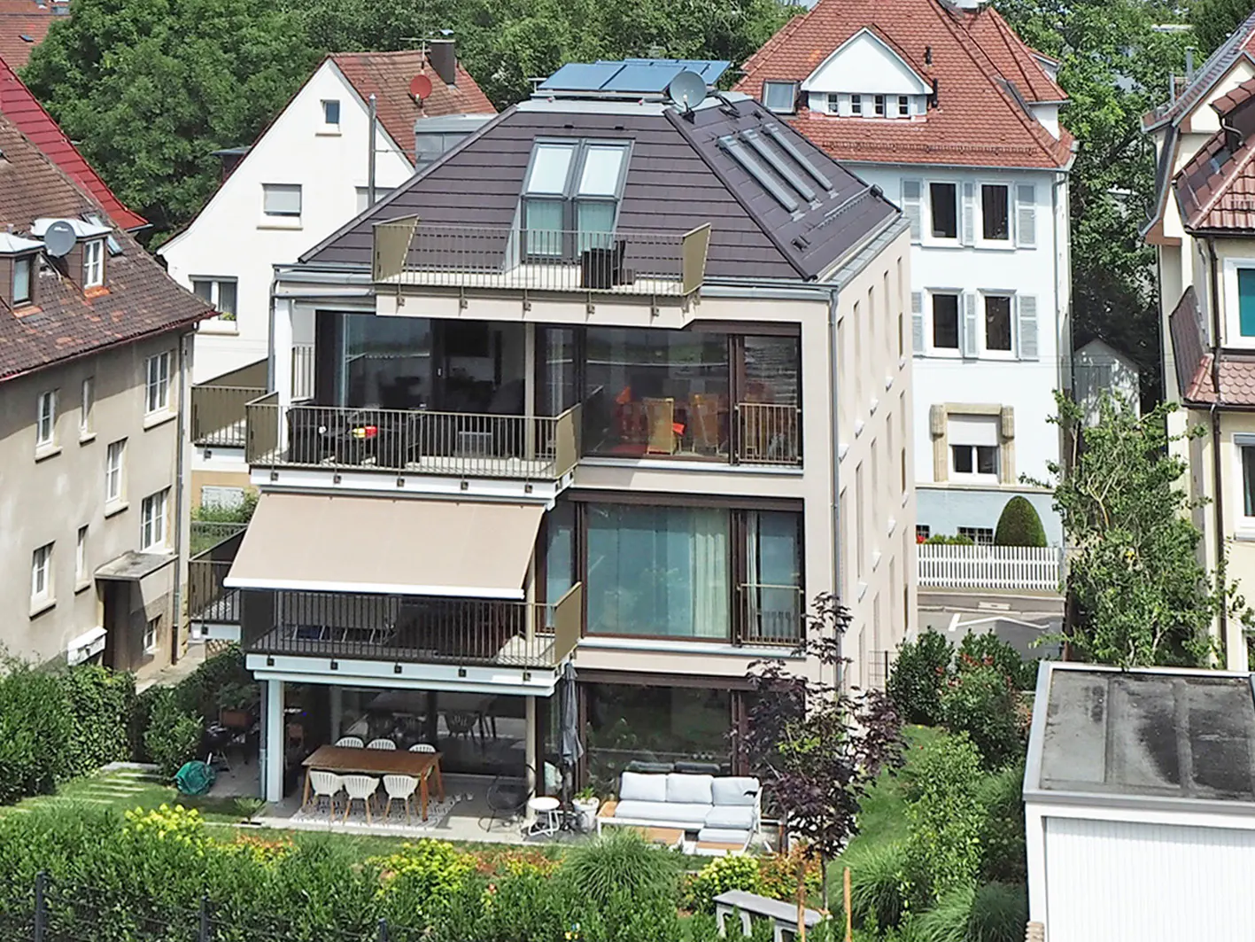 Multi-story modern home with balconies, glass walls, and a brown tile roof. Awnings shade the balconies. A garden is in the foreground.