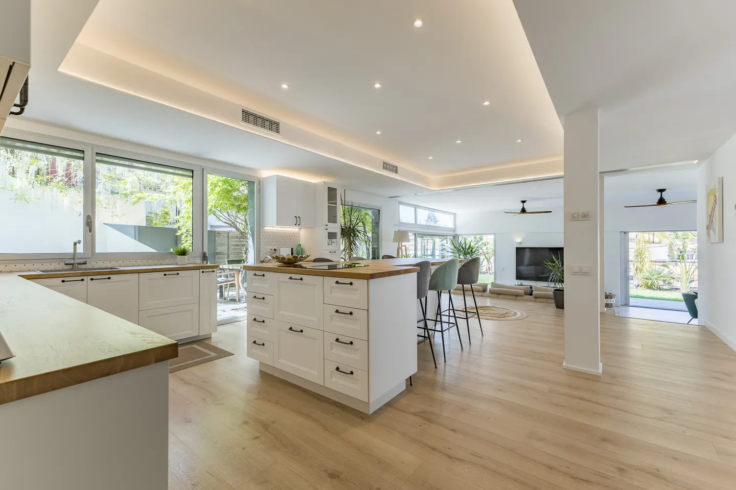 Bright, open-concept kitchen and living area with white cabinets, wood countertops, and light wood floors. Bar stools at the island. Large windows and doors to the outside.
