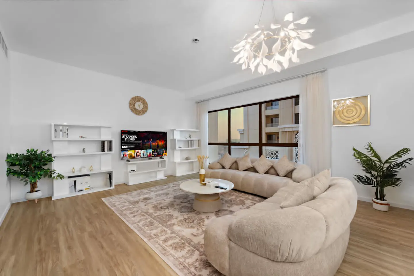 Bright living room with wood floors, white walls, and a beige sectional sofa. A TV displays "Stranger Things" above white shelves. A large window offers a city view.