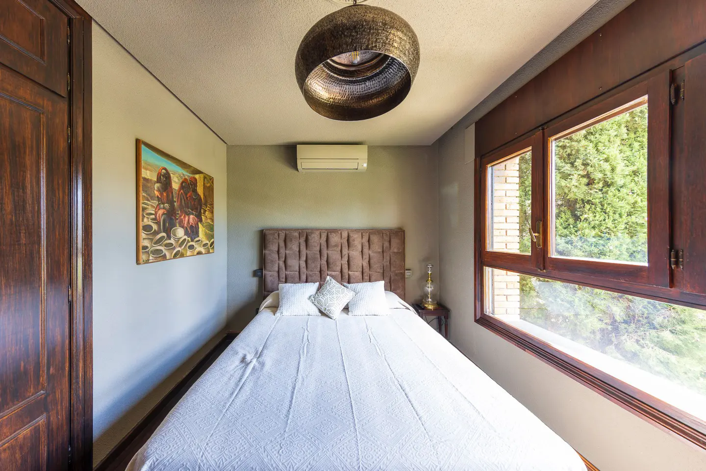 Bedroom with a white bed, brown headboard, and a large window with a view of green trees. A brown closet is on the left.