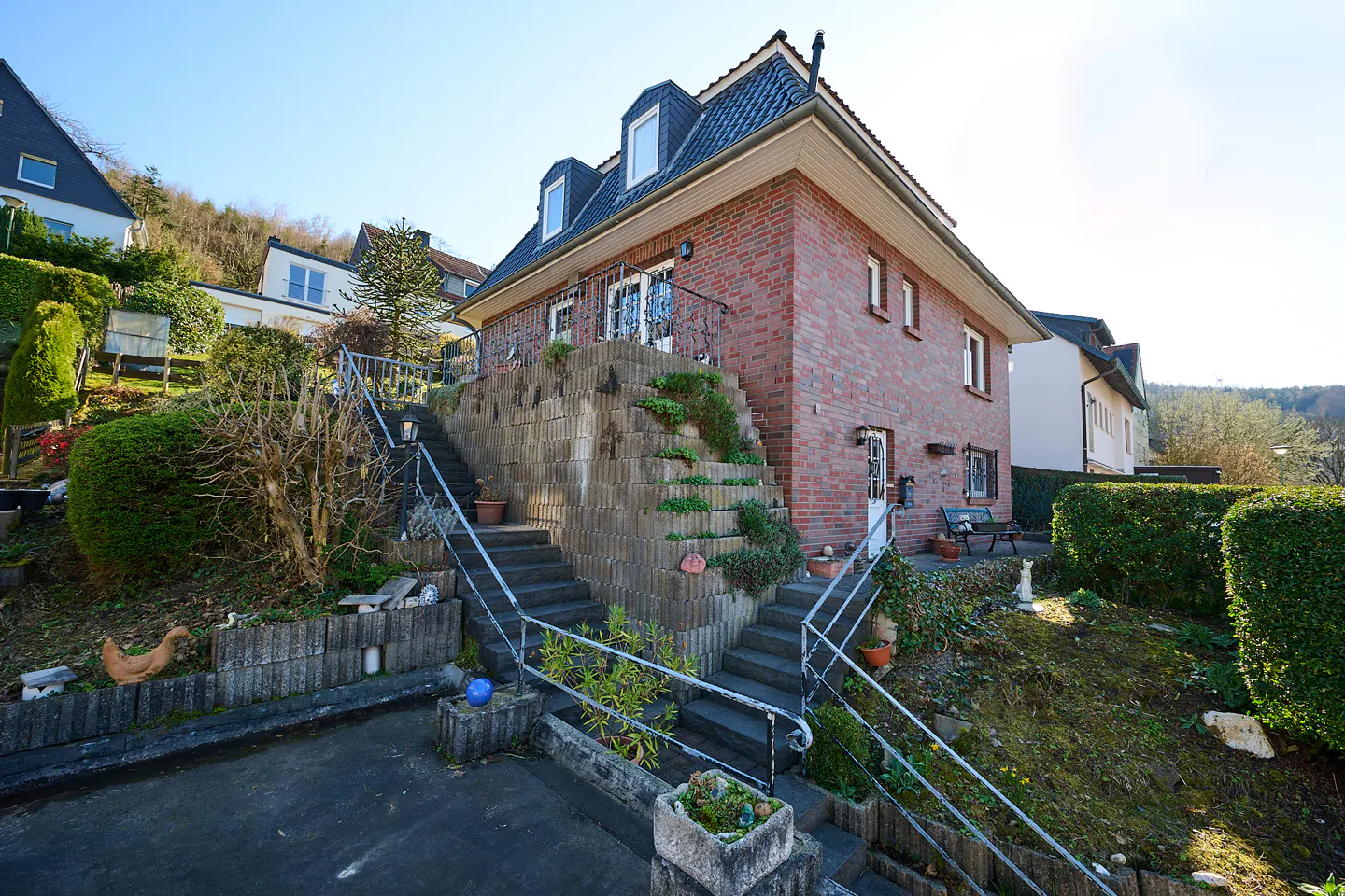 Exterior view of a two-story red brick house with a dark roof and dormer windows, accessed by two sets of stairs with metal railings.
