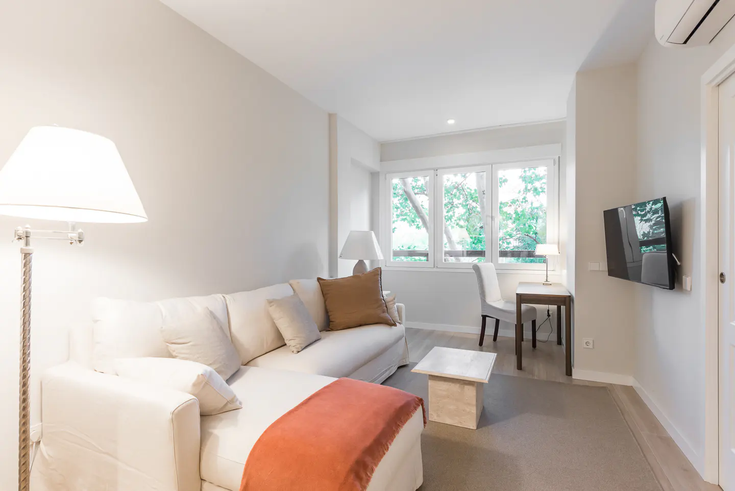 Bright living room with a white sofa, orange throw, desk, chair, and TV. Large windows overlook green trees.