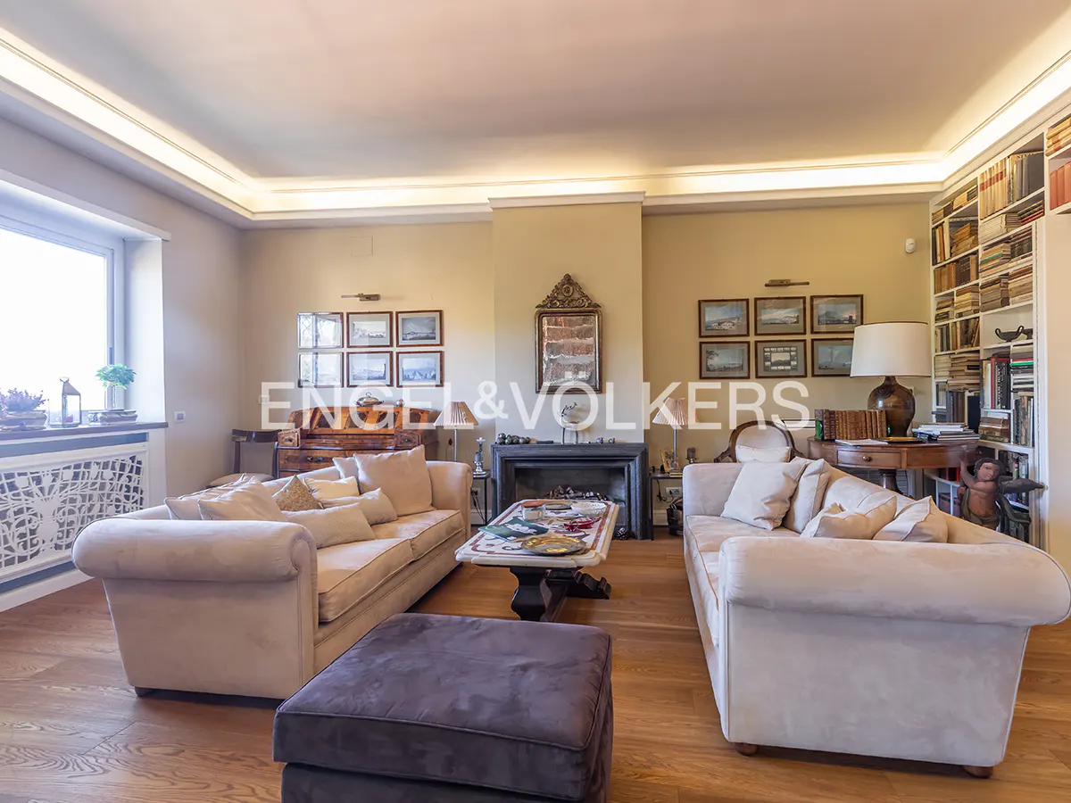 Living room with two sofas, a fireplace, and a bookshelf filled with books. The walls are beige and there are framed pictures on the walls.