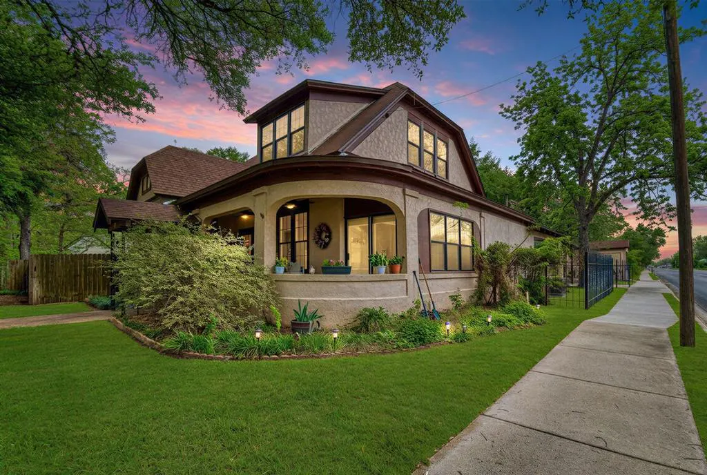 A two-story beige house with a brown roof, surrounded by green trees and grass at dusk.