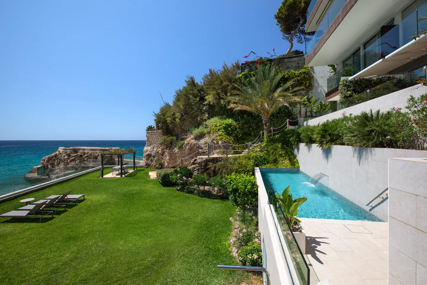 Luxury home with a pool overlooking the ocean. Green lawn, lounge chairs, and a pergola are visible.
