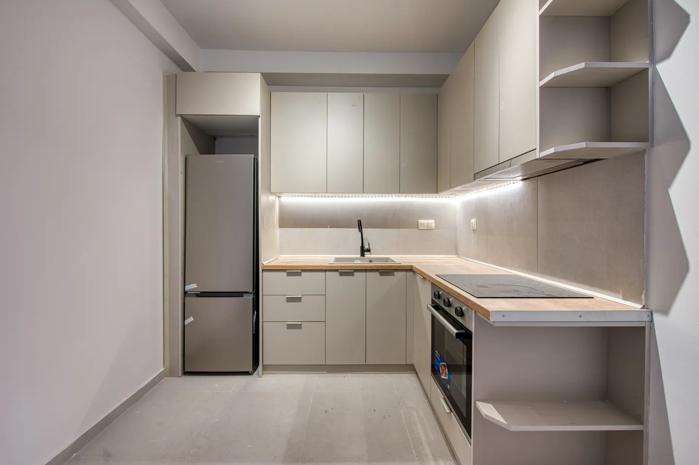 A modern kitchen with light gray cabinets, stainless steel refrigerator, and wood countertops. LED strip lighting illuminates the backsplash.