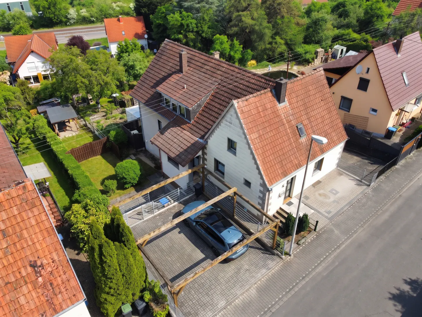 Aerial view of a white house with a red tile roof, featuring a wooden carport with a parked car and a lush green garden.