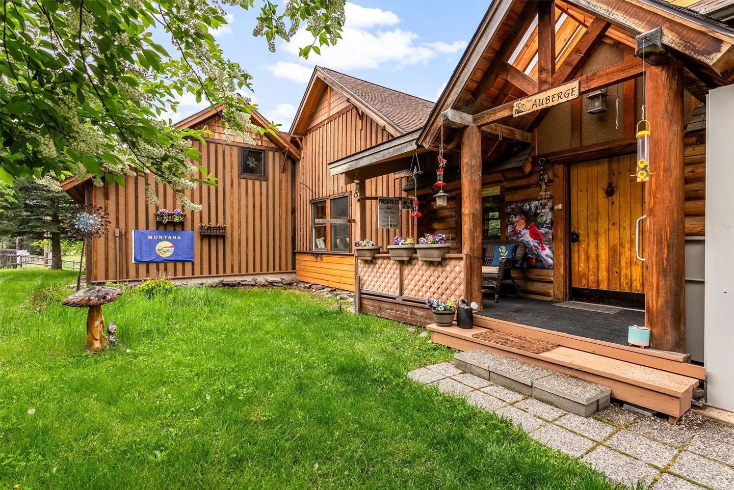 Exterior view of a rustic, wooden building with "So. 1st Auberge" sign, flower boxes, and a Montana flag on the lawn.
