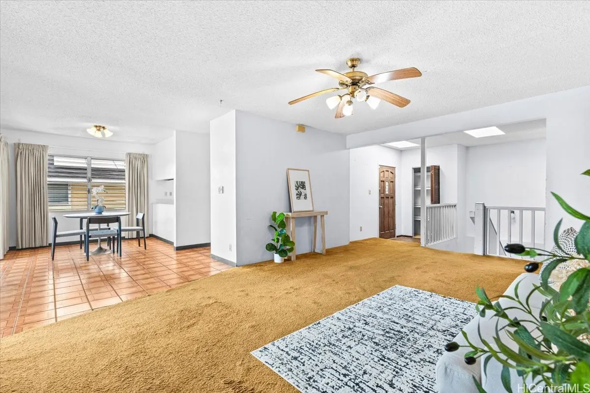 Open-concept living space with white walls, gold carpet, and tile flooring. A dining table, ceiling fan, and decor add to the room's charm.