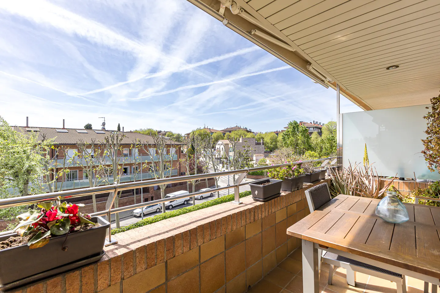 Balcony view with a wooden table, chairs, and potted plants. A street with buildings and trees is visible in the background under a blue sky.