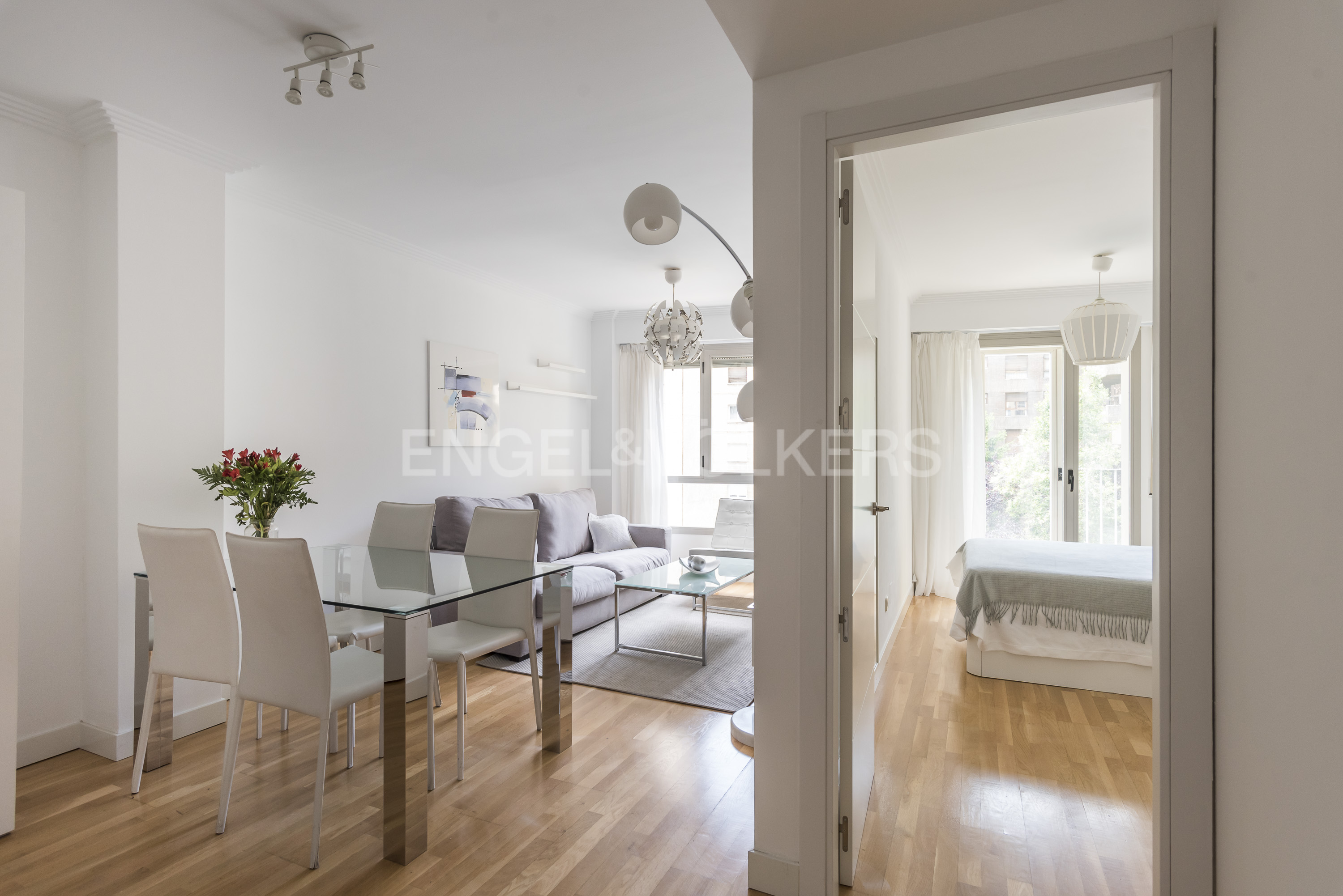 Bright living room with glass dining table, white chairs, and sofa, leading to a bedroom with hardwood floors. [AI-generated text]
