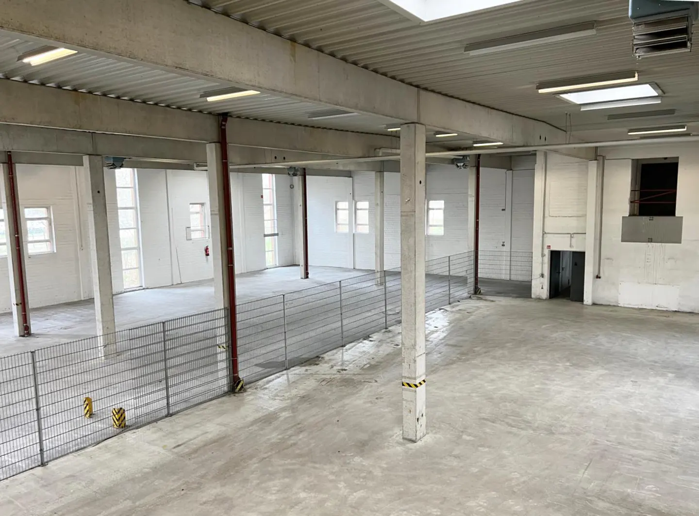 A large, empty warehouse with concrete floors, white walls, and a high ceiling with lights. Metal fencing divides the space.