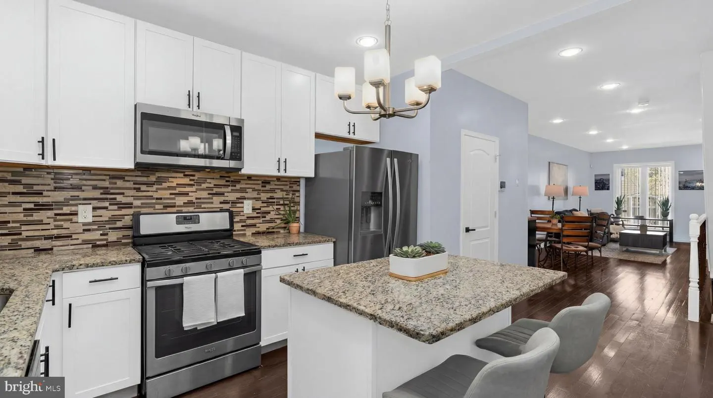 A modern kitchen with white cabinets, stainless steel appliances, and a granite countertop island with two gray chairs.