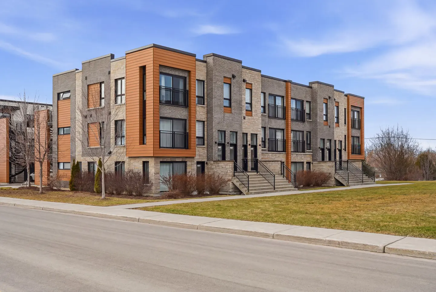 A modern, two-story townhouse complex with gray brick and wood-look siding under a blue sky. Black balconies and stair railings accent the facade.