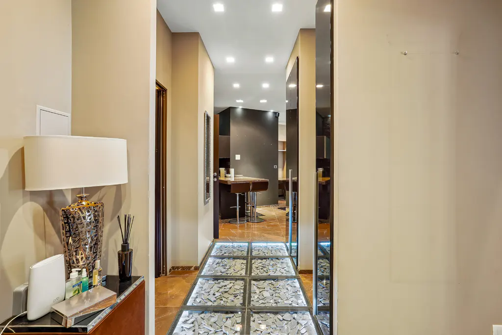 Hallway with a decorative lamp on a console table, leading to a kitchen with a stone floor and a glass-tiled walkway.