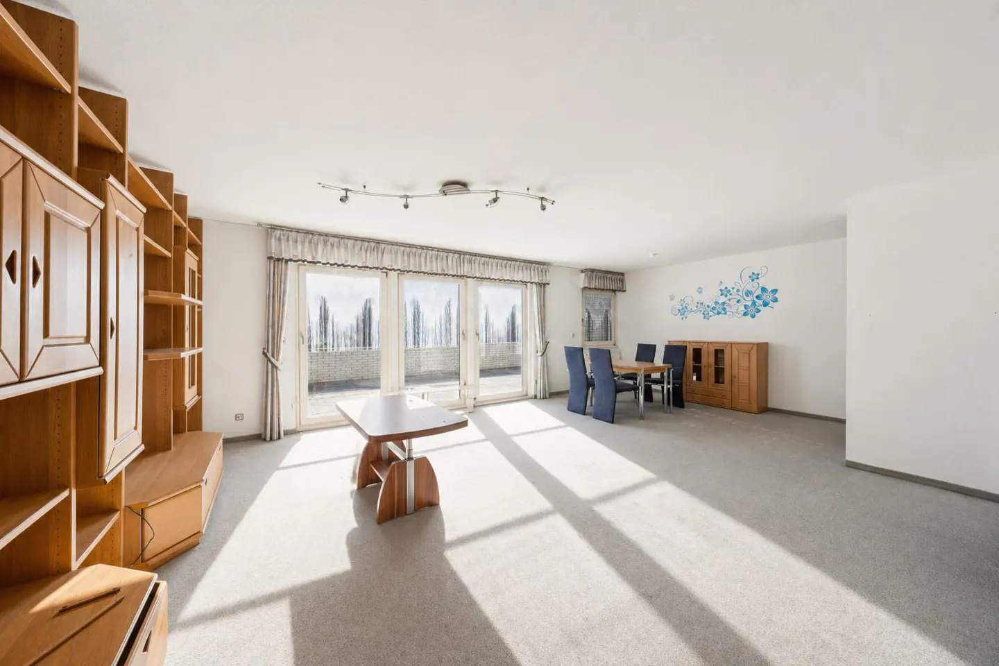 Bright living room with wood furniture, a dining table with blue chairs, and large windows leading to a balcony.