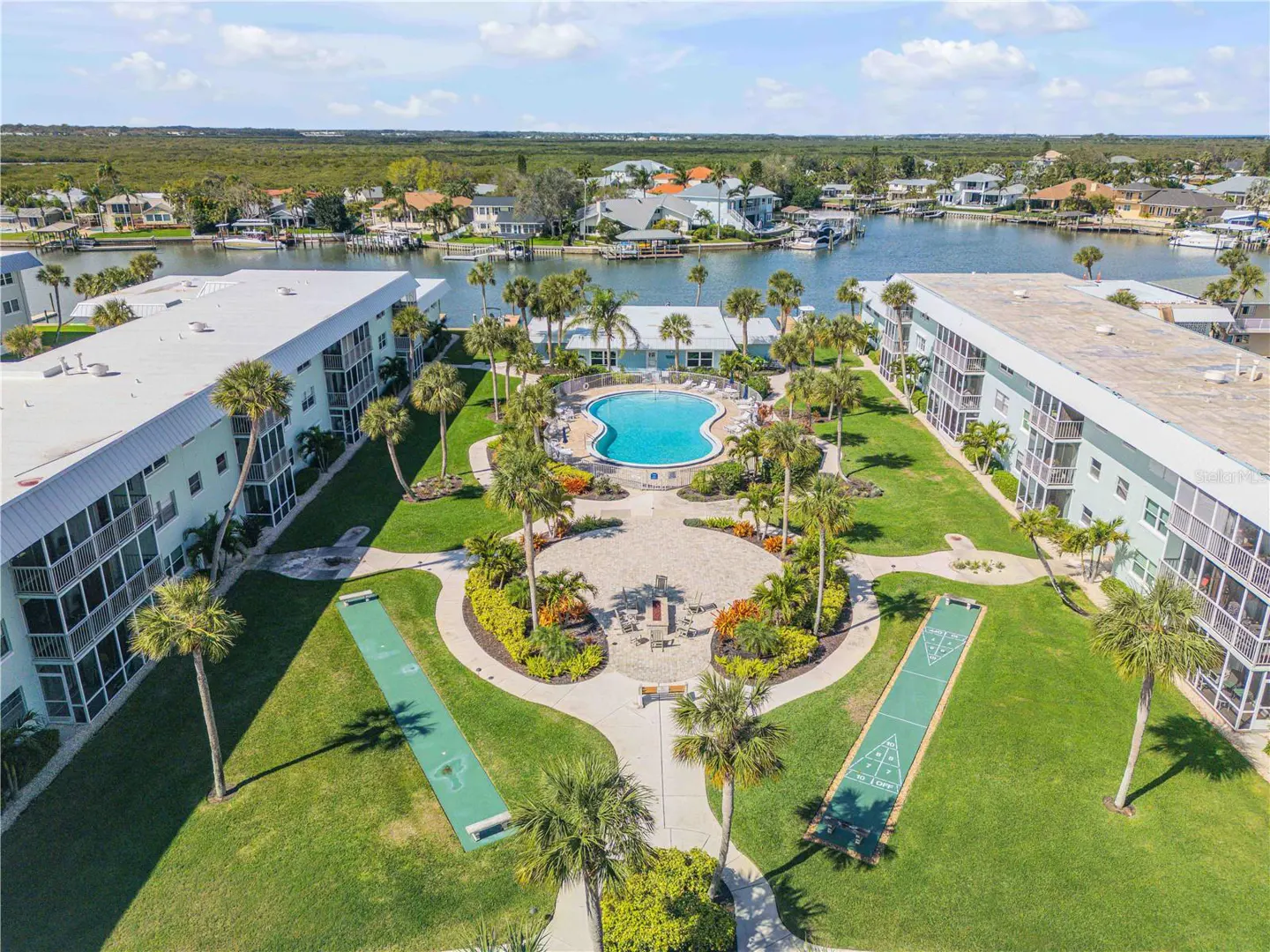 Aerial view of condo complex with pool, shuffleboard, palm trees, and waterfront access.