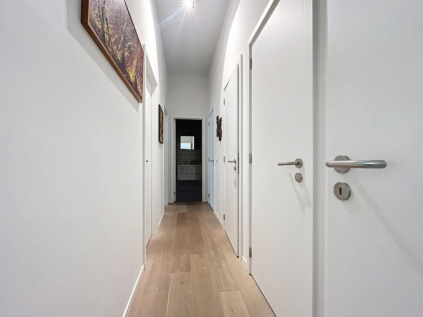 A bright hallway with white walls, light wood floors, and white doors. Artwork hangs on the left wall. A bathroom is visible at the end of the hall.