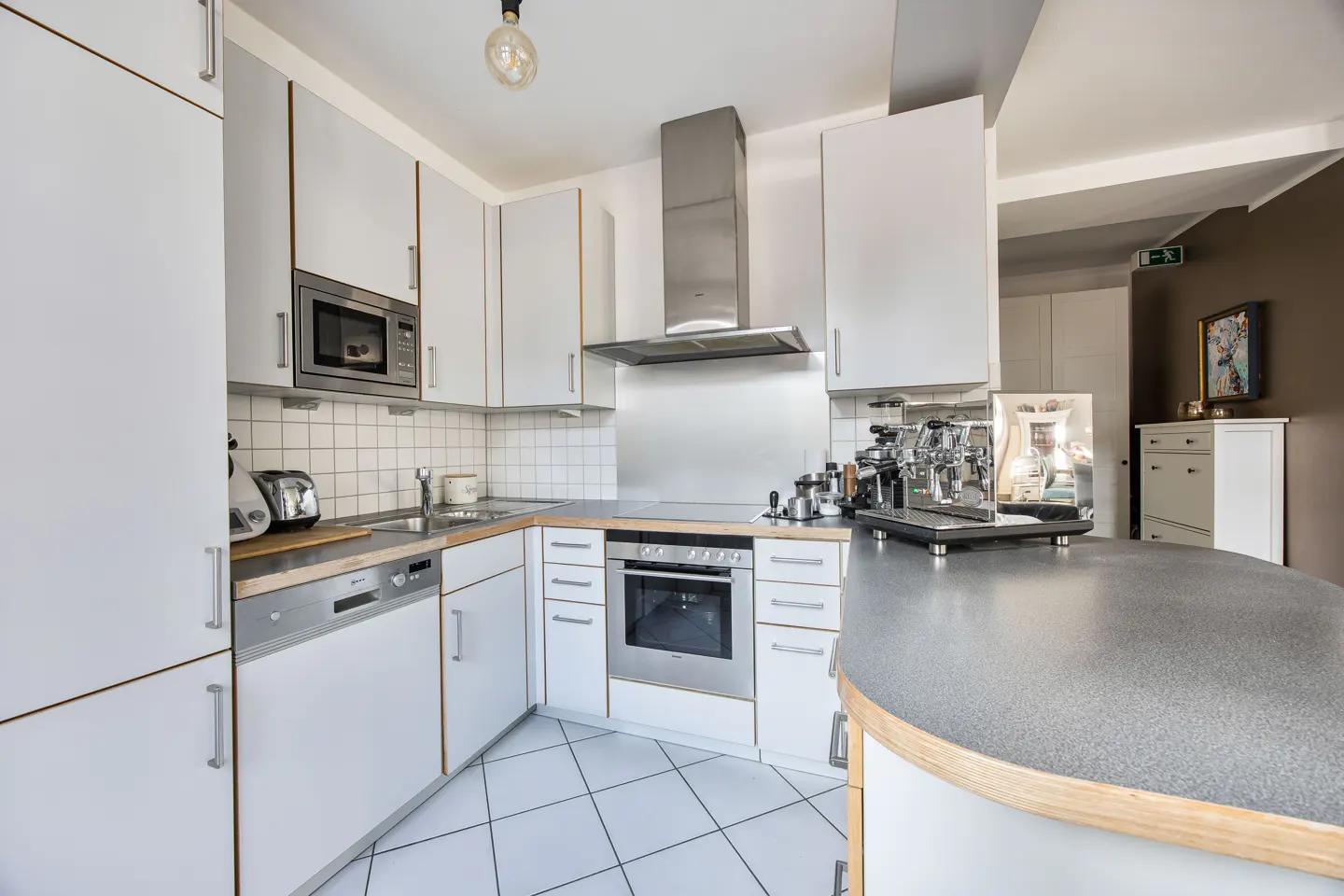 Bright kitchen with white cabinets, stainless steel appliances, and a gray countertop. White tiled floor and backsplash.