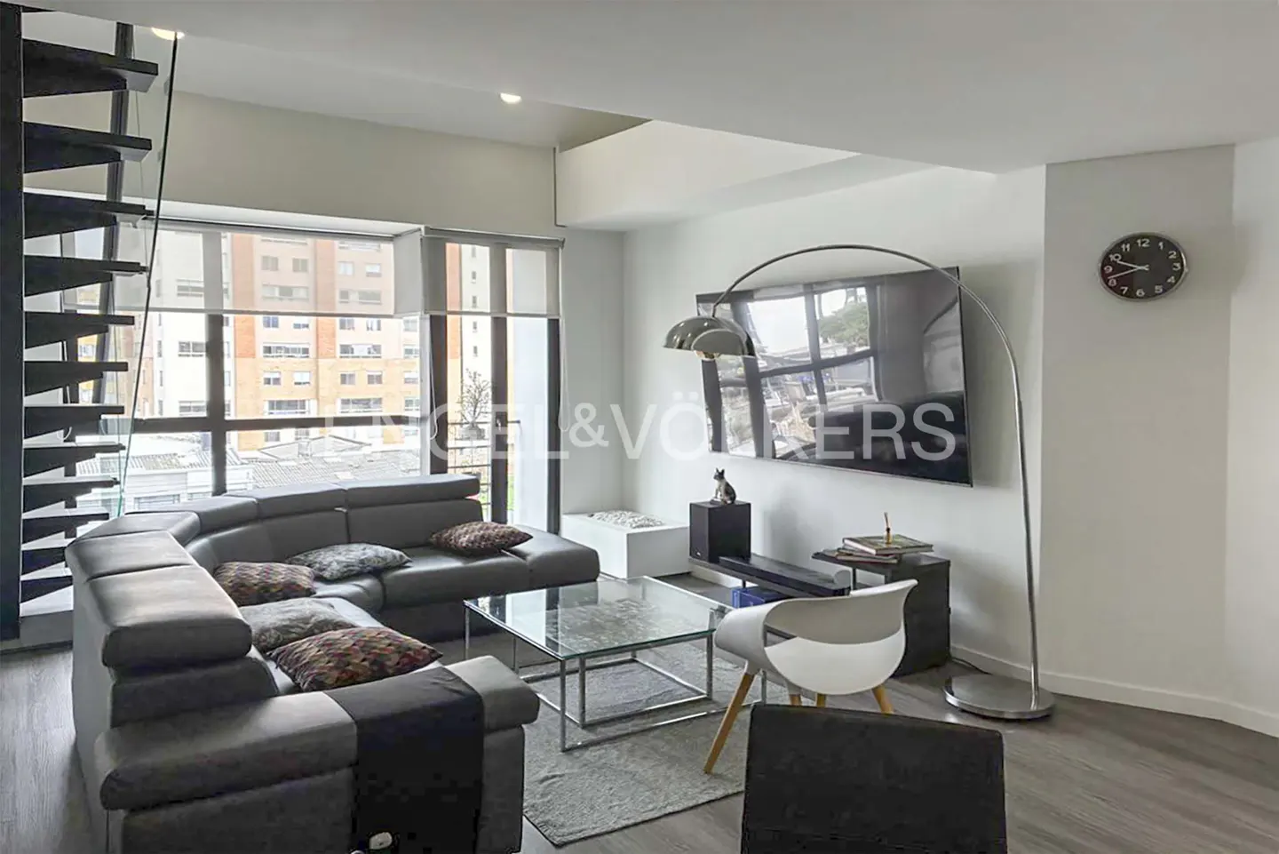 Living room with gray sectional sofa, glass table, TV, and modern floor lamp. Black spiral staircase on the left.