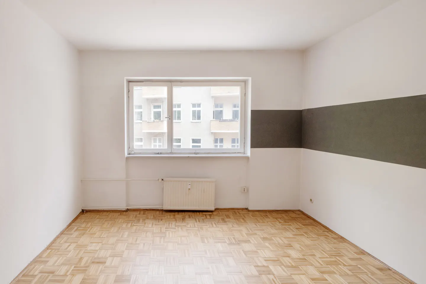 Empty room with parquet floor, white walls, and a gray stripe. A window shows a building across the street.