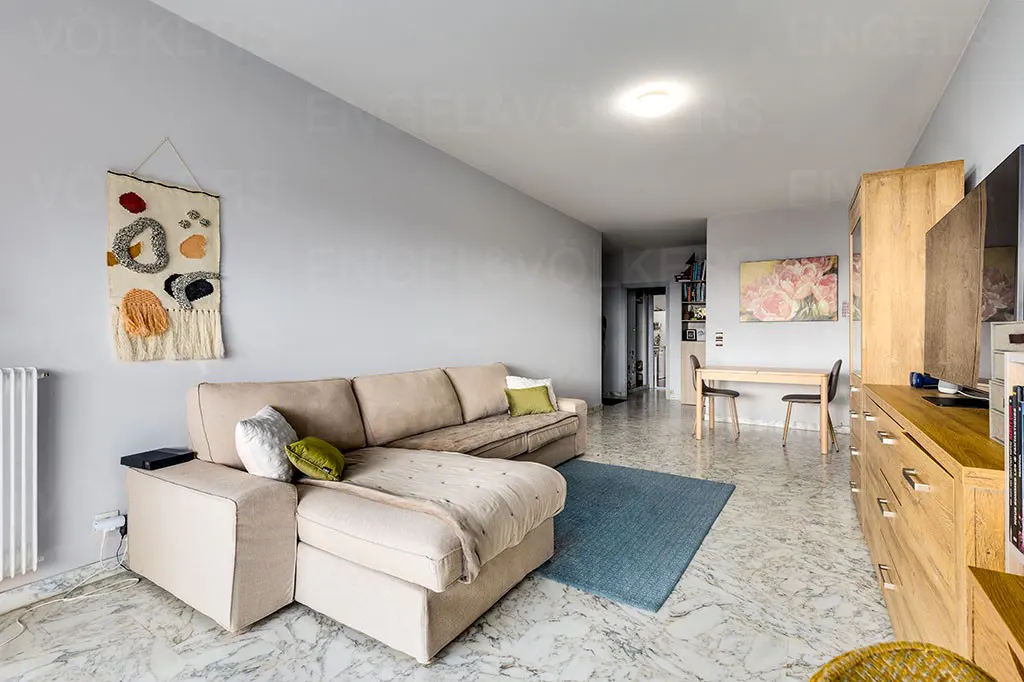 Living room with beige sectional sofa, blue rug, and light wood furniture on a marble floor. A tapestry hangs on the gray wall.