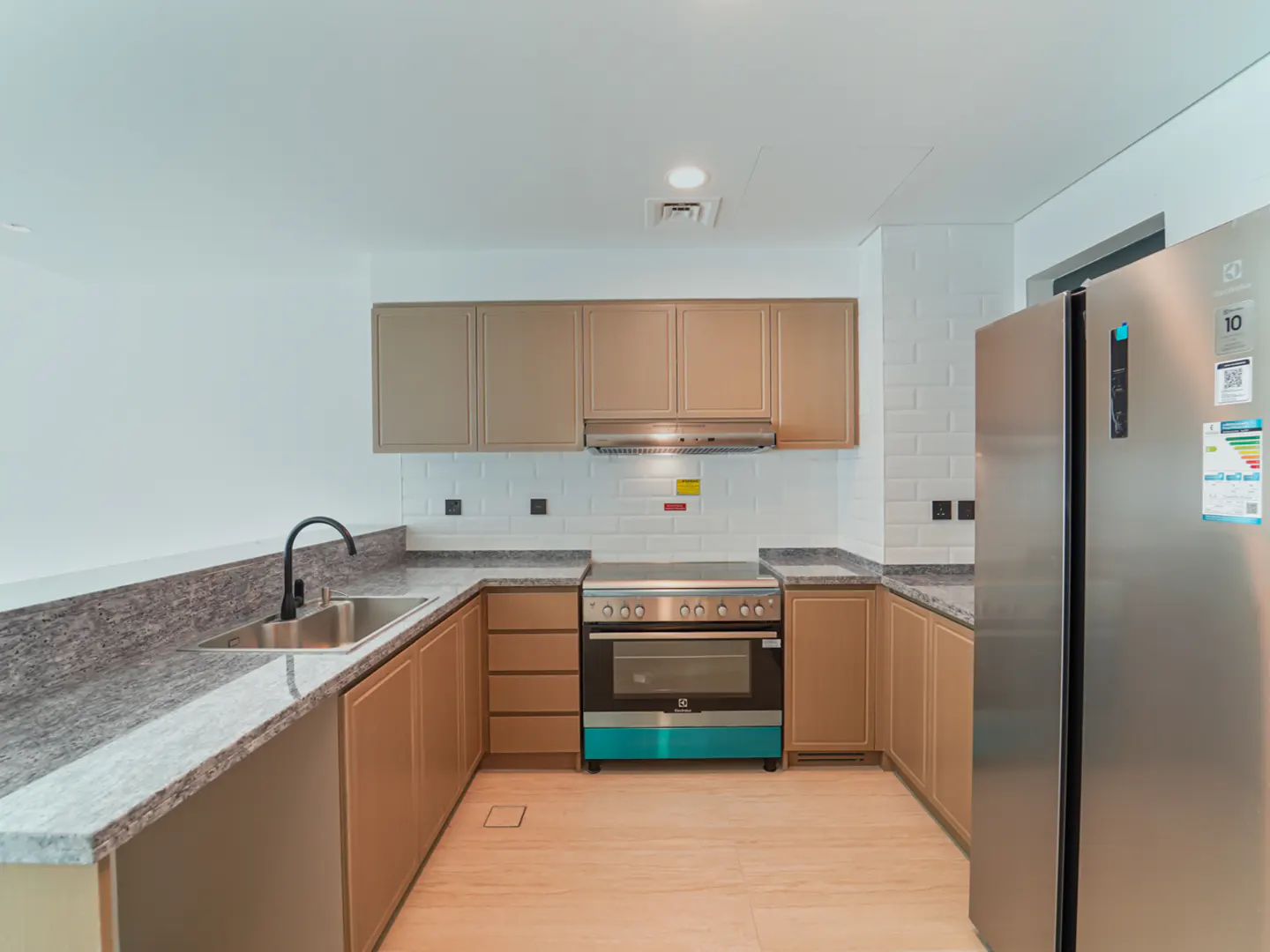A modern kitchen with tan cabinets, granite countertops, and stainless steel appliances. A sink with a black faucet is on the left, and a refrigerator is on the right.
