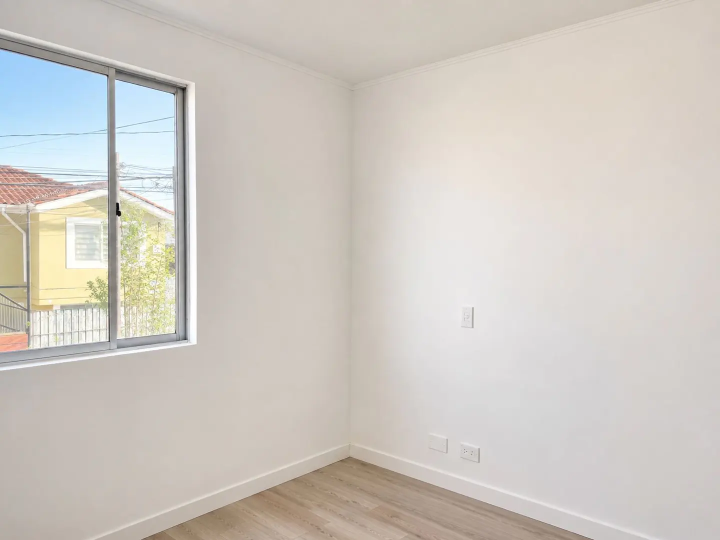 Bright, empty room with light wood floors and white walls. A window shows a yellow house and blue sky.