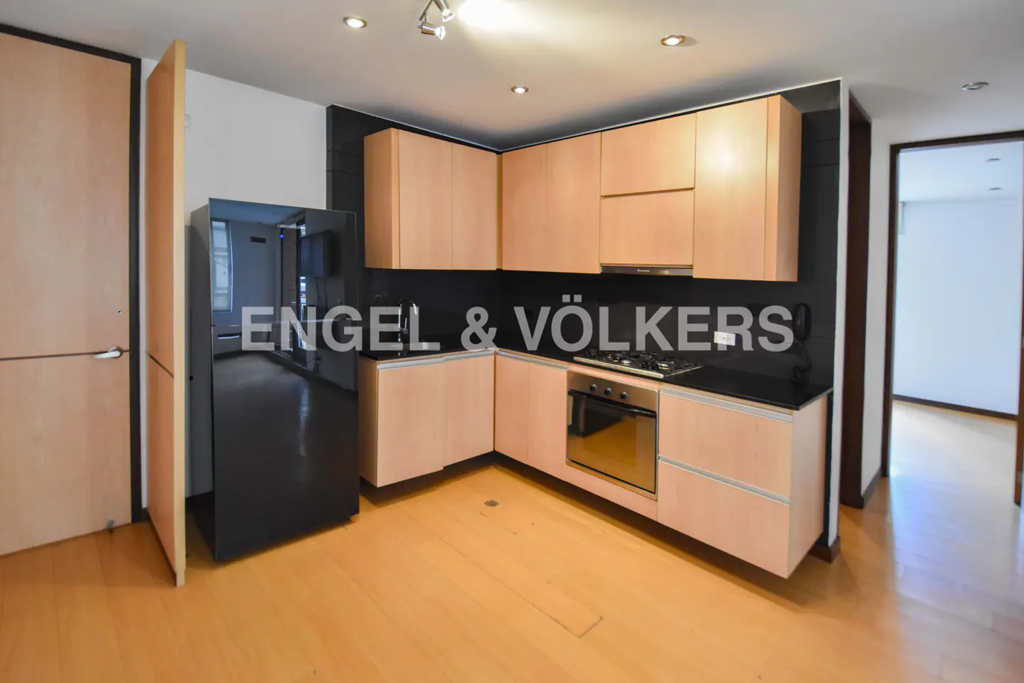 A modern kitchen with light wood cabinets, black countertops, and a stainless steel oven. A black refrigerator stands to the left.