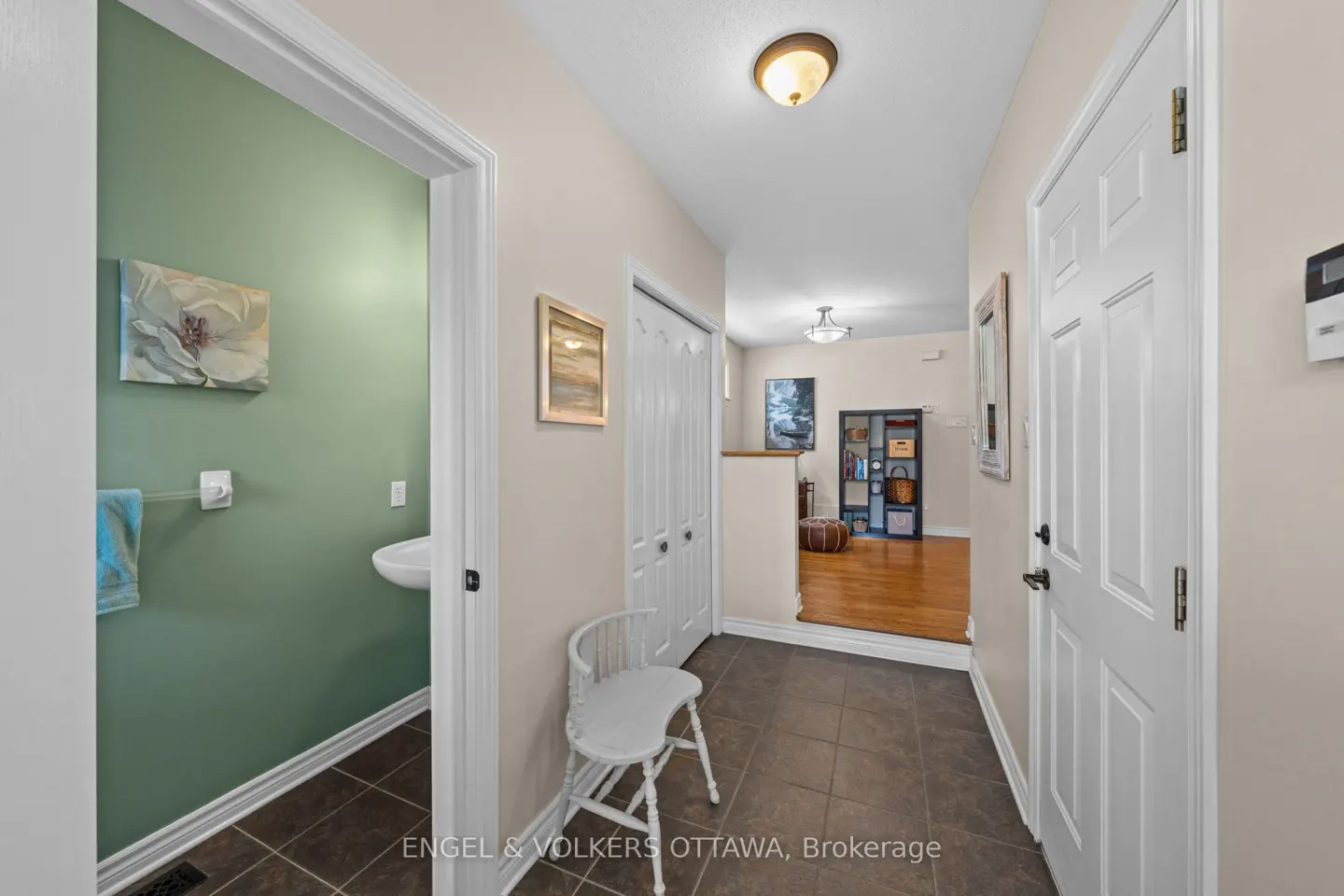 Hallway with brown tile floor, white walls, and a white door. A white chair sits near a closet. A bathroom is visible through a doorway.