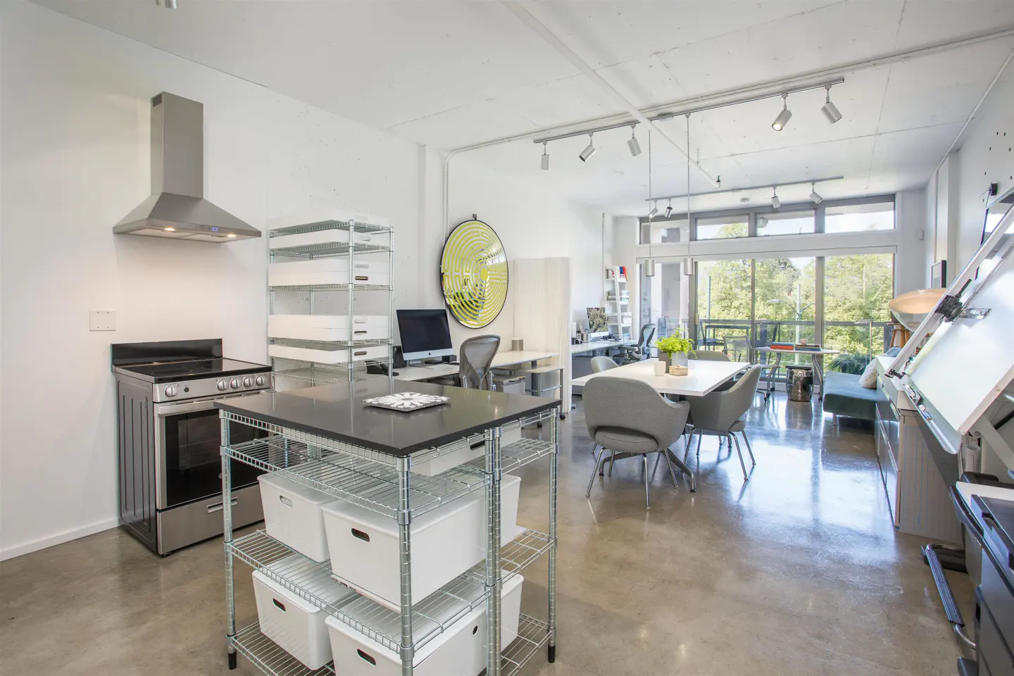 Open-concept loft with kitchen, dining, and office areas. Stainless steel appliances, wire shelving, and large windows with green trees outside.