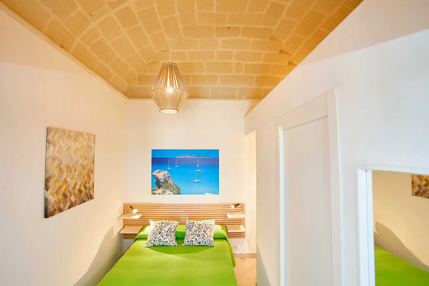Bright bedroom with a green bedspread, patterned pillows, and a beach scene picture above the headboard. A woven lamp hangs from the brick ceiling.