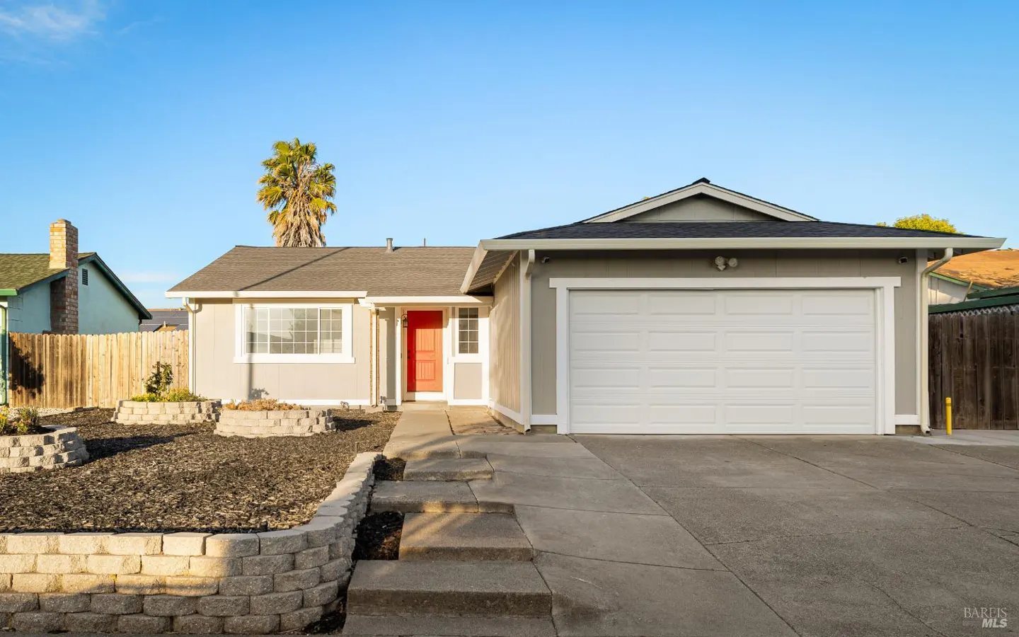 A single-story house with gray siding, a red front door, and a white garage door. A palm tree is visible in the background.