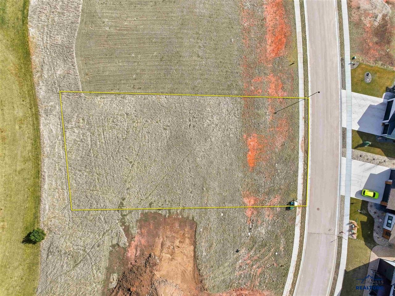 Aerial view of a vacant residential lot outlined in yellow, situated next to a paved road and nearby houses. [AI-generated text]
