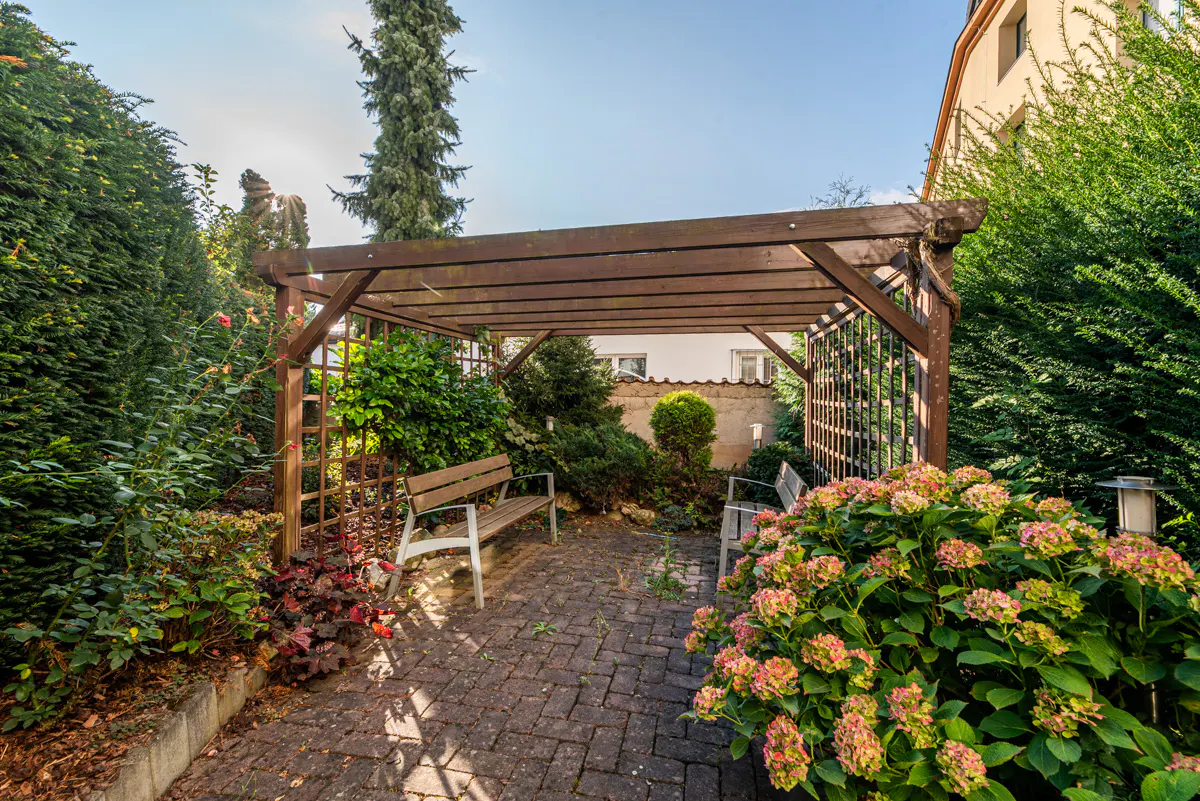 A wooden pergola covers a brick patio with two benches, surrounded by lush green hedges and blooming pink hydrangea bushes.