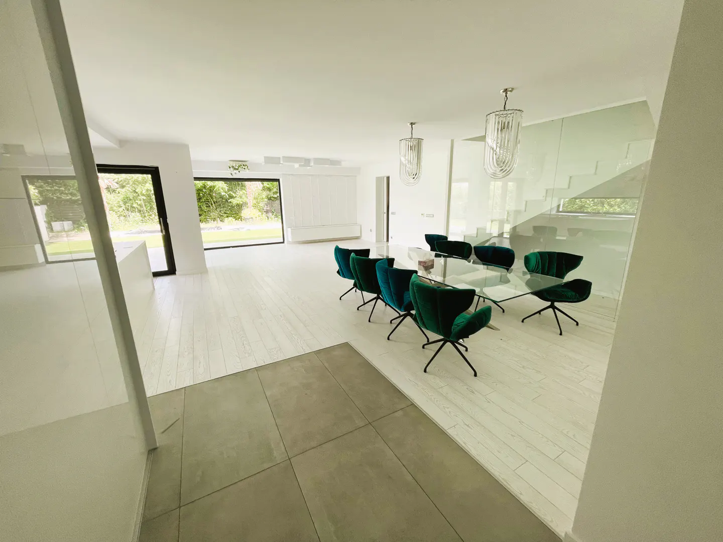 Bright, modern dining room with emerald chairs around a glass table. White walls, wood floors, and large windows offer a view of greenery.