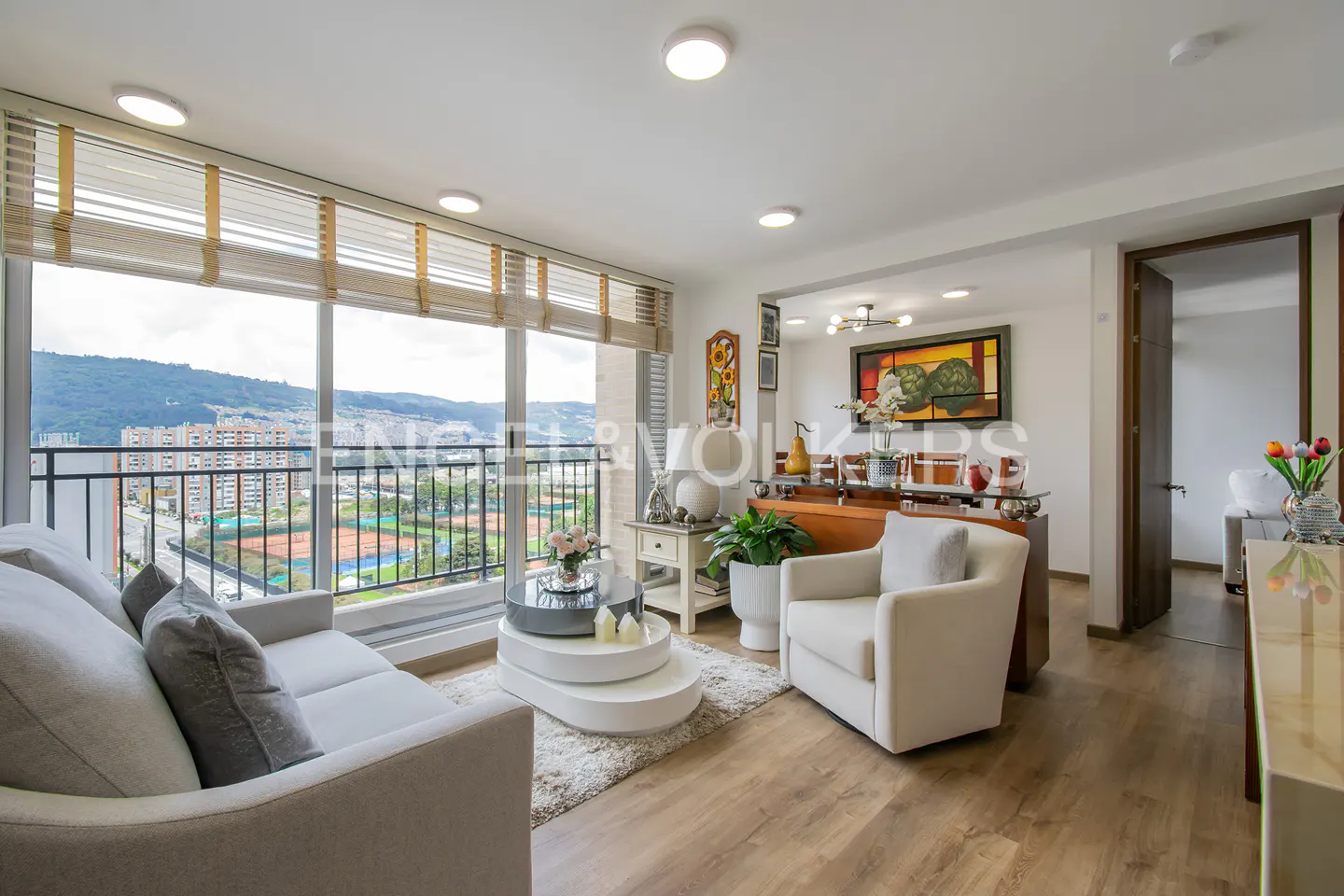 Bright living room with a gray sofa, white armchair, and tiered coffee table. Large windows offer a city view.