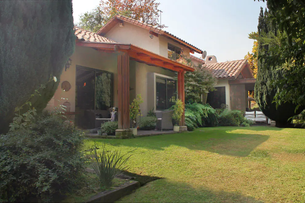 Exterior view of a beige house with a red tile roof and a green lawn. The house has a covered patio with wooden posts.