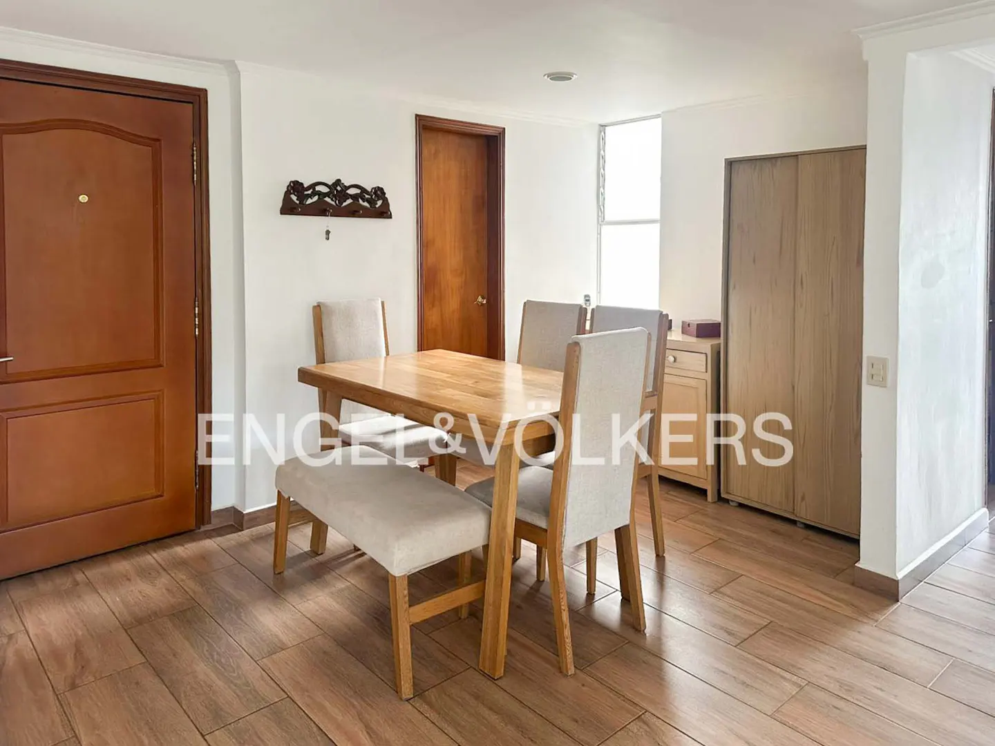 Dining area with a light wood table, a bench, and upholstered chairs on wood-look tile floors, near a wooden door and closet.