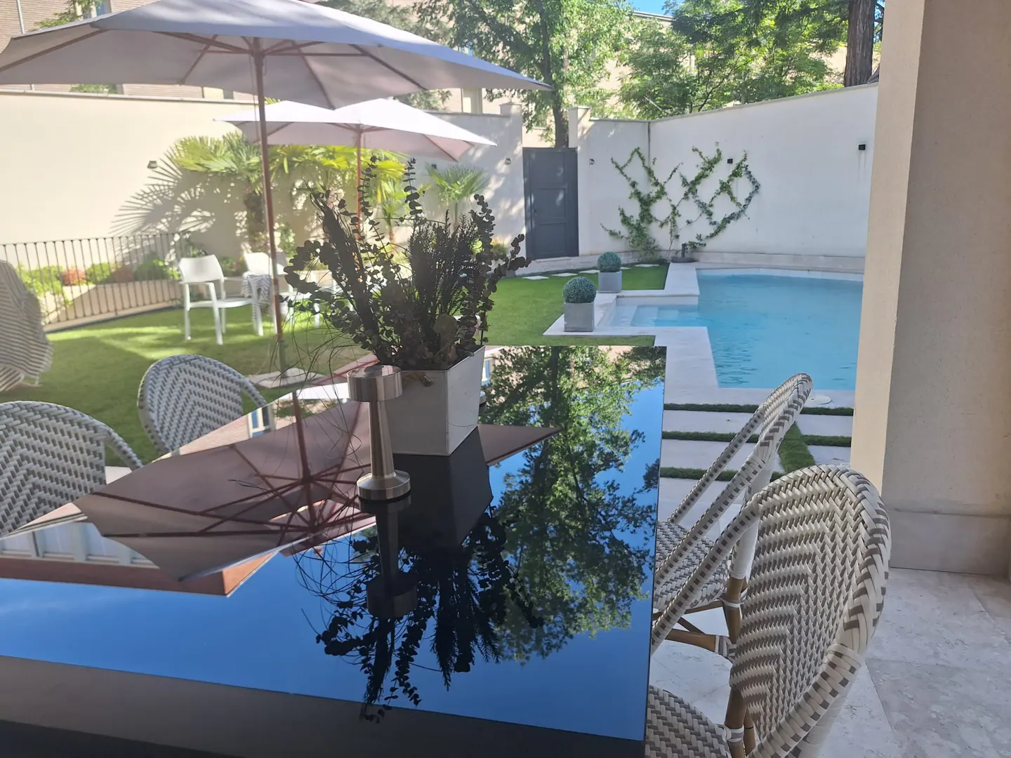 Outdoor patio with a black table, chairs, and a pool in the background. The table reflects the trees and sky.