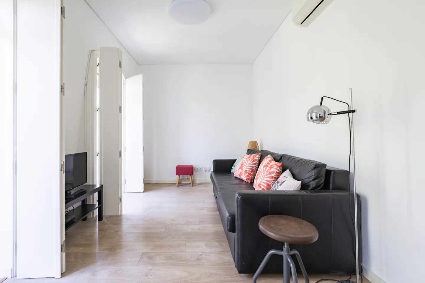 Bright living room with a black sofa, colorful pillows, a floor lamp, and a stool on a light wood floor. White walls and doors.