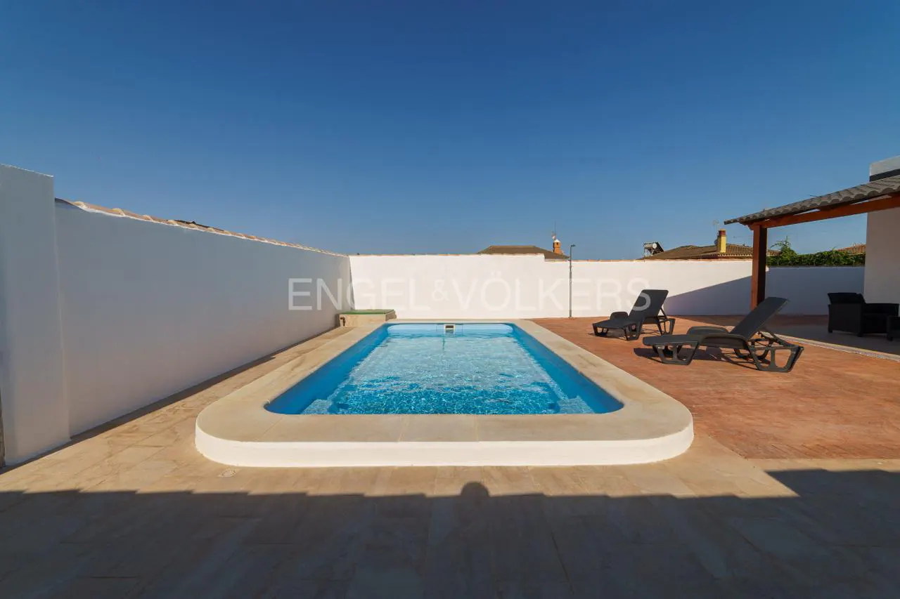 Outdoor pool with blue water, surrounded by a patio, white walls, and two black lounge chairs under a clear blue sky.