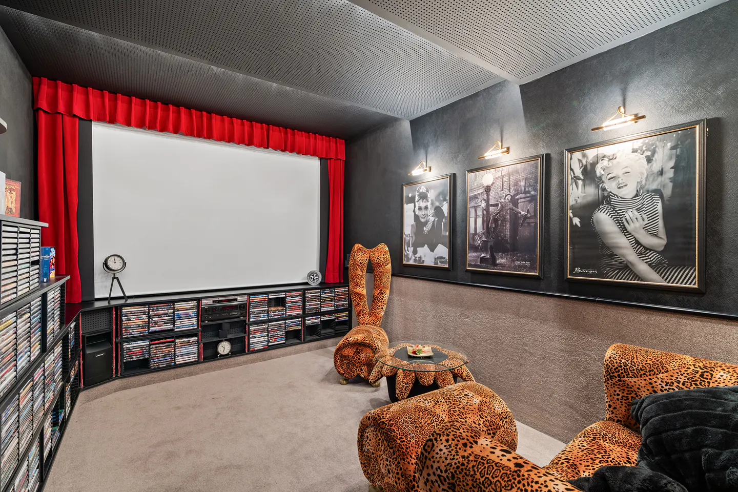 Home theater with a large screen, red curtains, leopard print chairs, and black and white portraits on a dark gray wall.