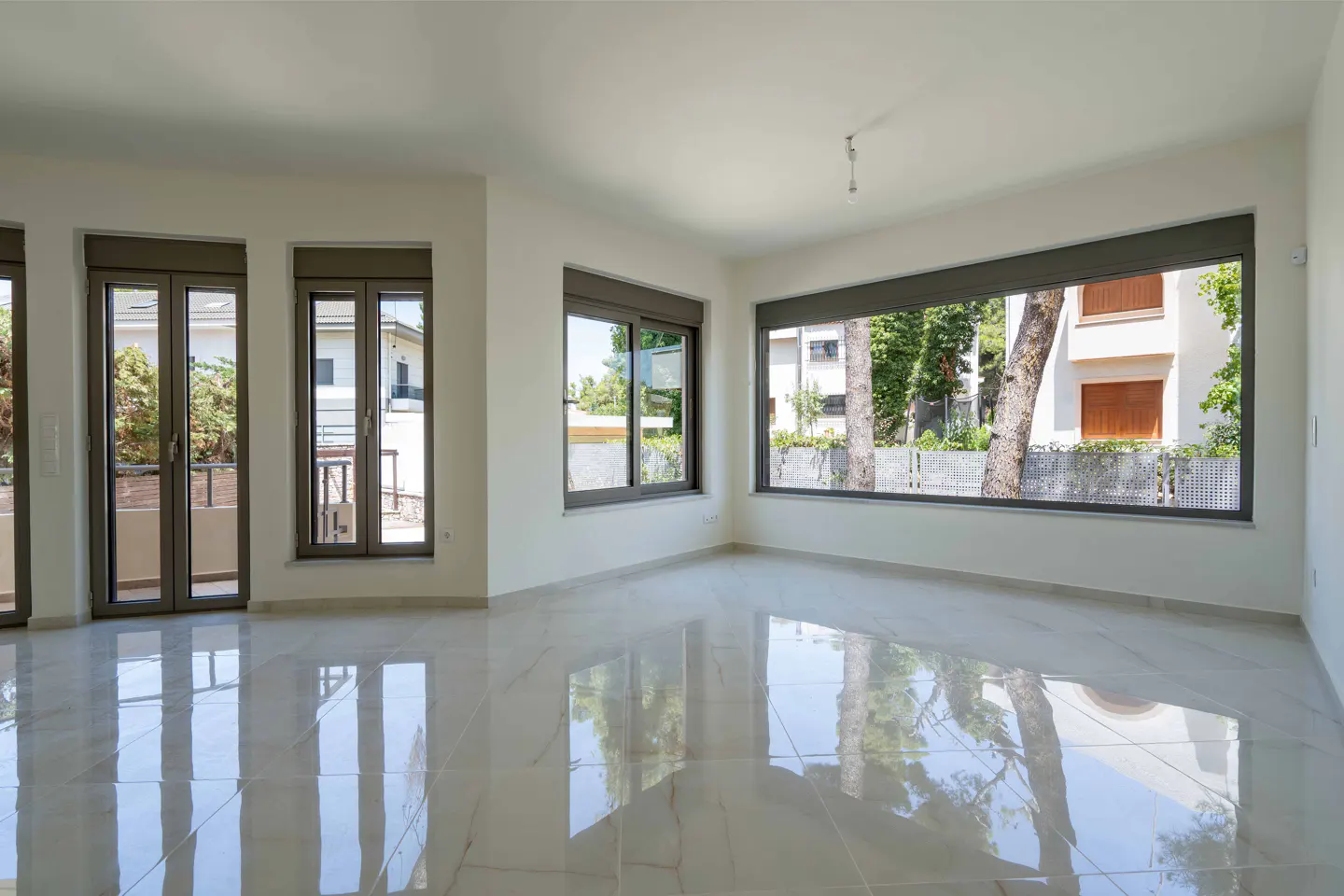 Bright, empty living room with white walls, polished marble floors, and large windows overlooking trees and houses.