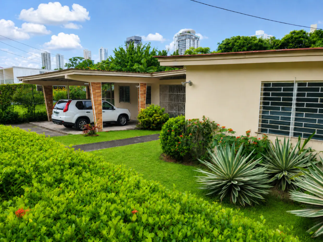 Beige single-story house with a carport, green lawn, and lush bushes. Two white cars are parked in the carport. Tall buildings are in the background.
