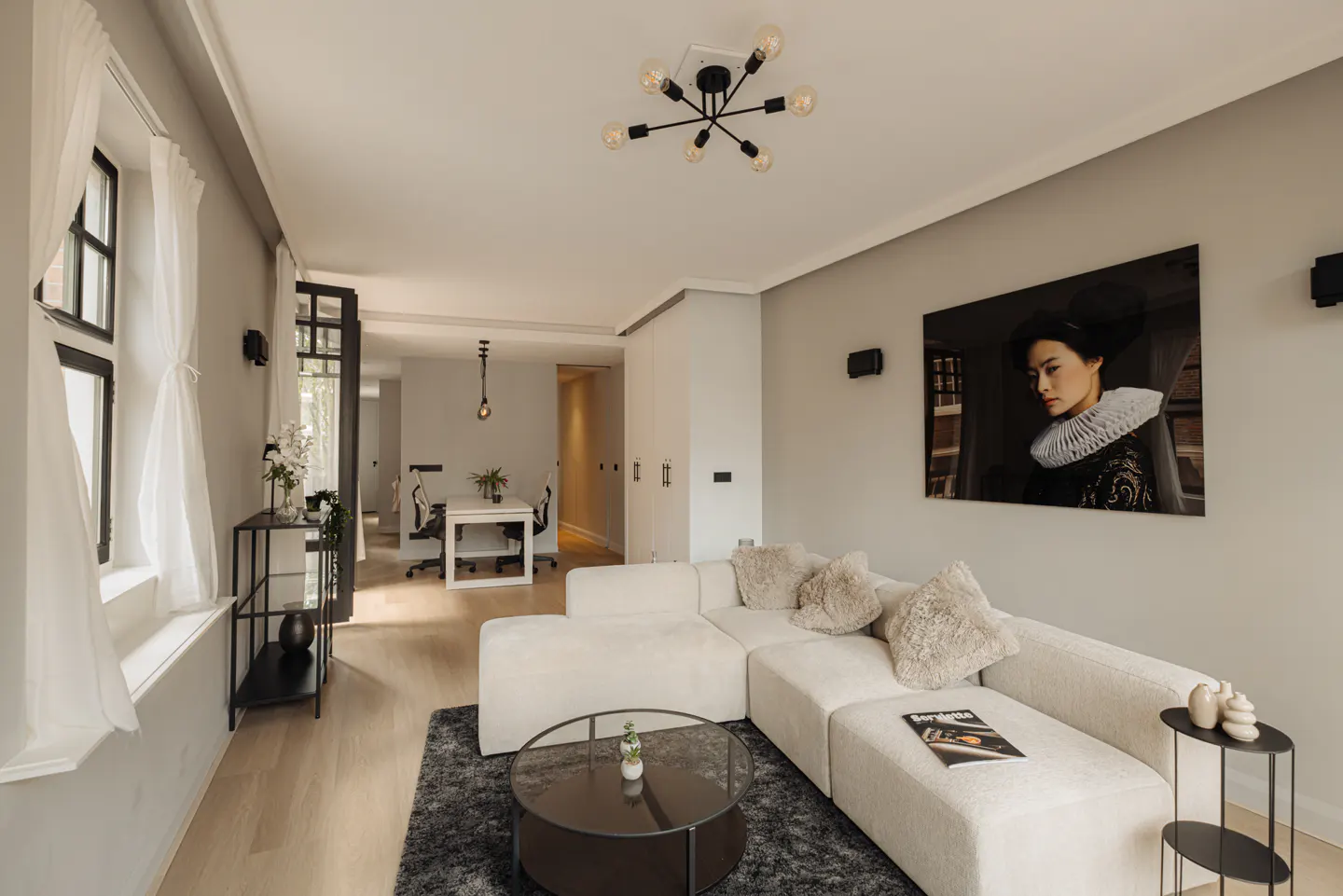Bright living room with a white sectional sofa, round glass coffee table, and a portrait on the wall. A black light fixture hangs from the ceiling.