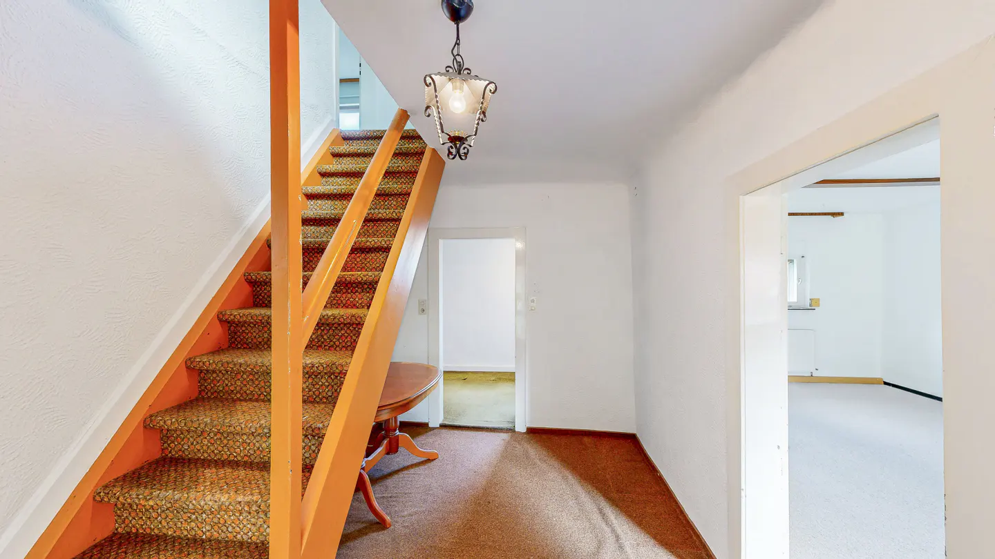 Entryway with orange wood stairs and patterned carpet. A vintage chandelier hangs from the white ceiling. Two doorways lead to other rooms.
