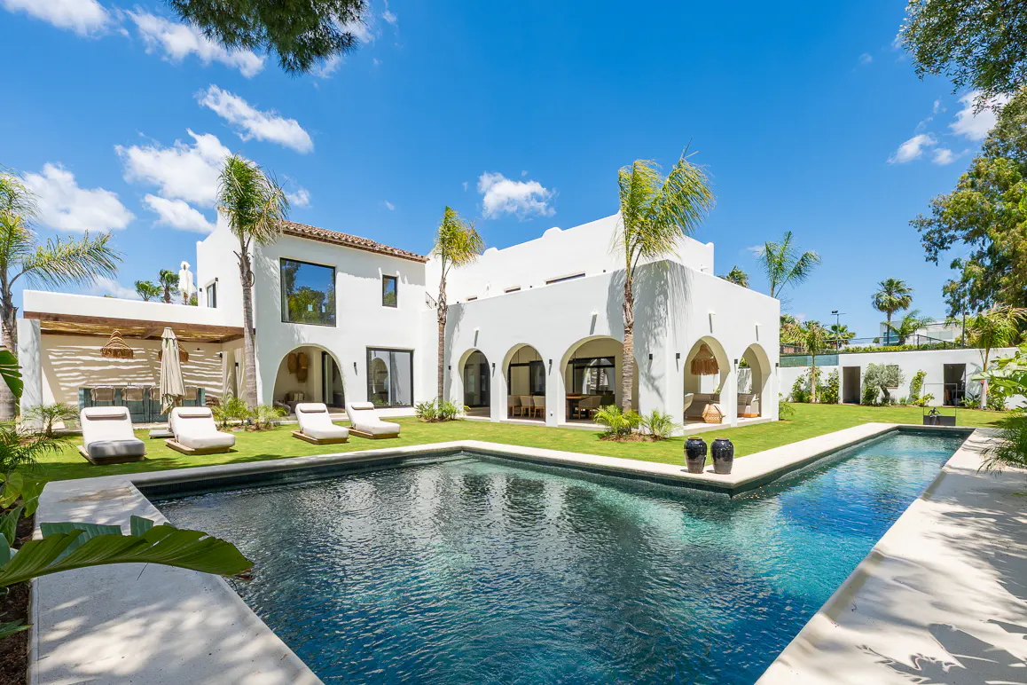 Exterior of a white, modern villa with a pool, lawn, and palm trees under a blue sky. Lounge chairs are on the grass.