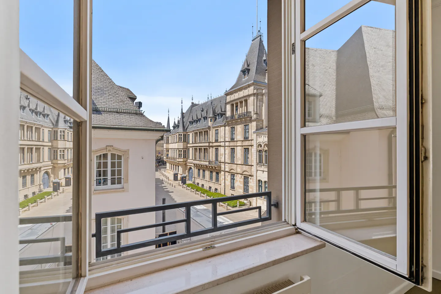 View from an open window overlooking a historic European street with ornate stone buildings and a clear blue sky.
